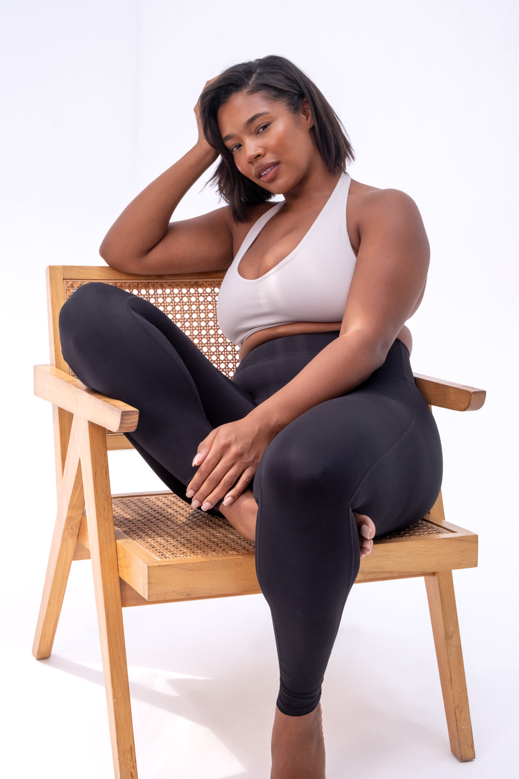 Woman sitting on a wooden chair wearing a white top and black leggings against a white background